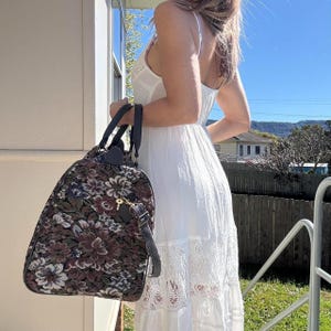 Könnte beinhalten: Eine dunkelblumige Tapisserie-Reisetasche mit schwarzen Ledergriffen und einem abnehmbaren Schultergurt. Die Tasche hat einen Schlüsselanhänger und einen Reißverschluss. Die Tasche wird von einer Person getragen, die ein weißes Kleid trägt.