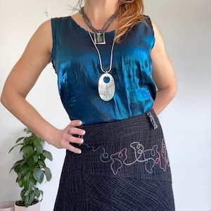 May include: A woman wearing a teal blue satin sleeveless top with a silver pendant necklace and a black skirt with colourful embroidery.