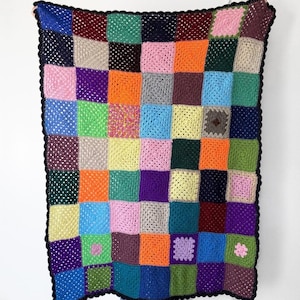 May include: A vibrant, handmade crochet blanket with a patchwork design. The blanket features squares in various colours, including blue, pink, orange, green, and purple, with a black border. The blanket is held up against a white background.
