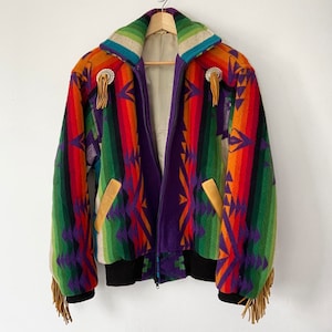 Puede incluir: Una chaqueta colorida de estilo suroeste con cierre de cremallera y detalles de flecos. La chaqueta presenta un patrón geométrico en tonos de rojo, naranja, amarillo, verde, azul y morado.