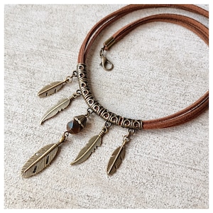 May include: A brown suede choker necklace with bronze-colored feather pendants and a central geometric bead. The necklace features a decorative metal bar and a clasp closure, showcasing a bohemian style.