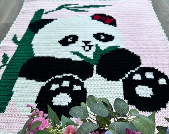 May include: A crocheted blanket with a panda bear design. The panda is black and white with a red bow, set against a pink background. Green bamboo stalks frame the panda. The blanket has a scalloped edge.
