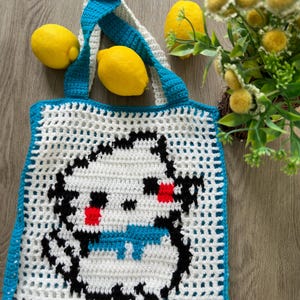 May include: A white crochet tote bag featuring a pixelated character design in black, red, and blue yarn. The bag has a grid-like open weave pattern, a solid blue border, and two blue crochet handles. The design depicts a stylized creature.