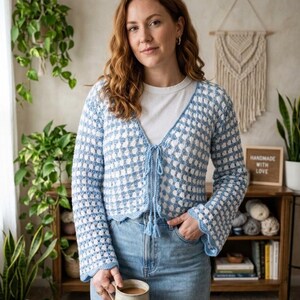May include: A light blue and white crochet cardigan with a checkered pattern. The cardigan has a V-neck, bell sleeves, and a tie closure. The image also shows a white t-shirt, blue jeans, and a mug. A sign reads "HANDMADE WITH LOVE."