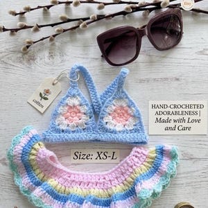May include: A handmade crochet bikini set in pastel colors, including a light blue top with a floral design and a ruffled bottom. The set is labeled "Size: XS-L" and the tag says "cotton". Sunglasses and a sprig of pussy willow are also in the image.
