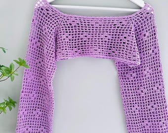 May include: A light purple, open-weave crochet shrug with long sleeves. The garment is displayed on a white hanger against a light gray background. The crochet pattern creates a grid-like texture. The sleeves are long and extend down from the cropped body.