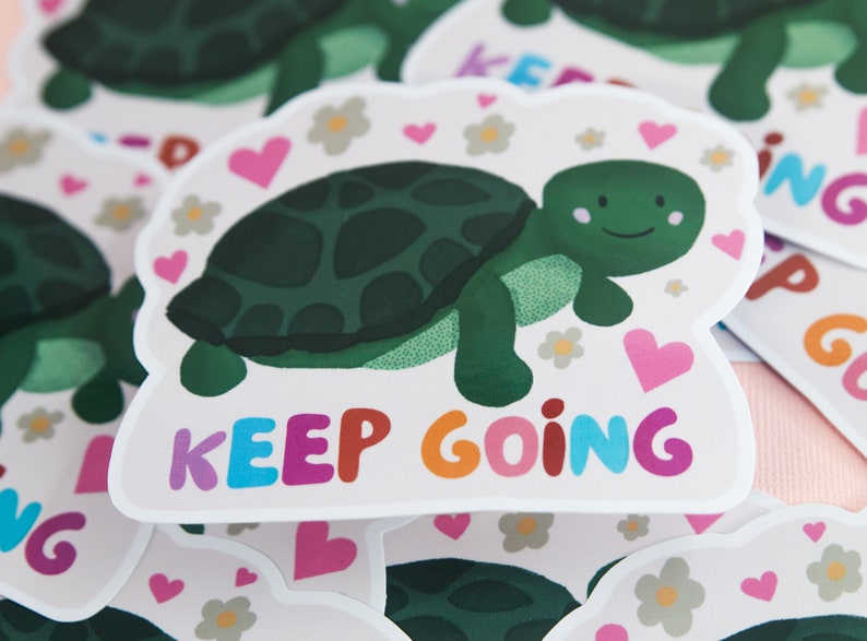 Pu&ograve; includere: Un adesivo bianco con una tartaruga verde e il testo "KEEP GOING" in lettere colorate. La tartaruga ha un viso sorridente ed &egrave; circondata da cuori rosa e fiori bianchi.