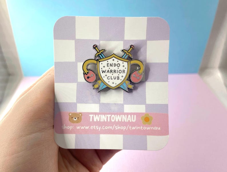 May include: A yellow and blue enamel pin with a shield and swords. The shield has the text "ENDO WARRIOR CLUB." The pin is on a white and purple checkered background.