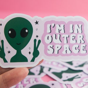 May include: A green alien with a peace sign on each hand, with a pink background and the text "I'm in outer space" in white.