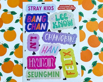 May include: A sheet of colorful stickers with the names of the members of the K-pop group Stray Kids. The stickers are on a white background with a pattern of orange citrus fruit.