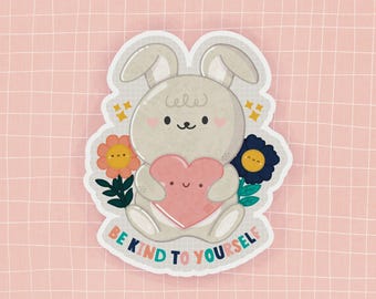 May include: A sticker featuring a cartoon bunny holding a pink heart, surrounded by flowers. The text "BE KIND TO YOURSELF" is written in a rainbow font. The background is a pink grid.