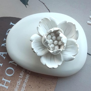 May include: A white, oval decorative stone with a detailed, three-dimensional white flower on top. The flower has multiple petals and a textured centre. The stone is smooth and appears to be made of ceramic or a similar material.