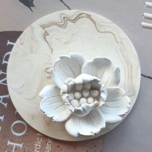 May include: A white, three-dimensional flower sculpture with detailed petals and a textured center, mounted on a round, cream-coloured base with a marbled pattern. The sculpture is likely made of ceramic or a similar material.
