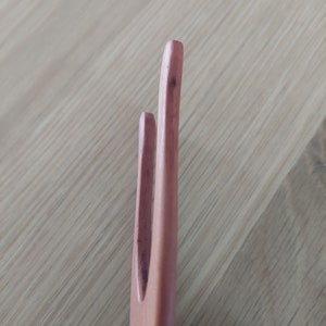May include: Close-up of a wooden fork with two tines. The fork is a light brown color and appears to be made of wood. The tines are rounded at the tips. The background is a light brown wooden surface.