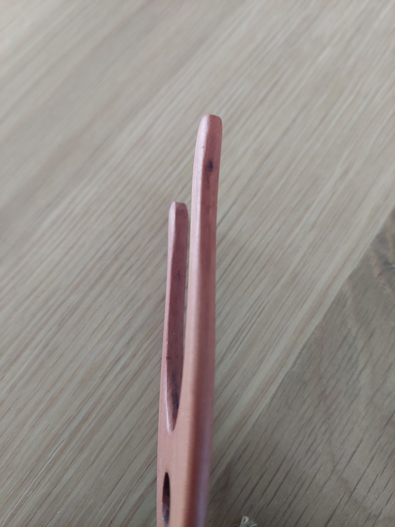 May include: Close-up of a wooden fork with two tines. The fork is a light brown color and appears to be made of wood. The tines are rounded at the tips. The background is a light brown wooden surface.
