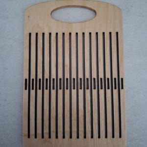 May include: A rectangular light-colored wooden weaving loom featuring a series of vertical laser-cut slots and a horizontal row of smaller rectangular cutouts across the center. The top edge includes a rounded handle cutout for easy gripping.