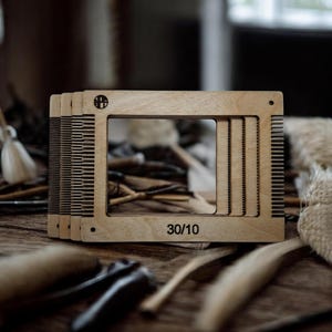 Peut inclure: Plusieurs outils de tissage en bois, avec un cadre rectangulaire et des dents en forme de peigne. Le texte "30/10" est visible sur le cadre. Les outils sont en bois clair et sont conçus pour les travaux textiles.