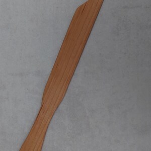 Könnte beinhalten: Ein Holzspatel mit einem langen, schlanken Griff und einer flachen, abgewinkelten Klinge. Der Spatel ist aus hellem Holz gefertigt, mit sichtbaren Maserungsmustern. Am Ende des Griffs ist ein kleines Loch gebohrt. Ideal zum Kochen und Backen.