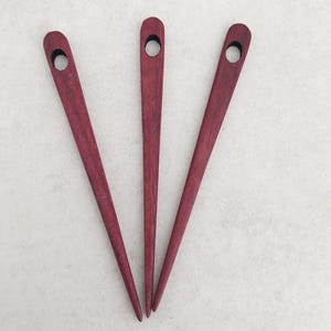 Könnte beinhalten: Drei Haarstäbe aus Holz in tiefem Burgunderrot. Jeder Stab hat ein spitzes Ende und eine abgerundete Oberseite mit einem kleinen Loch. Die Stäbe sind fächerförmig vor einem hellen Hintergrund angeordnet.