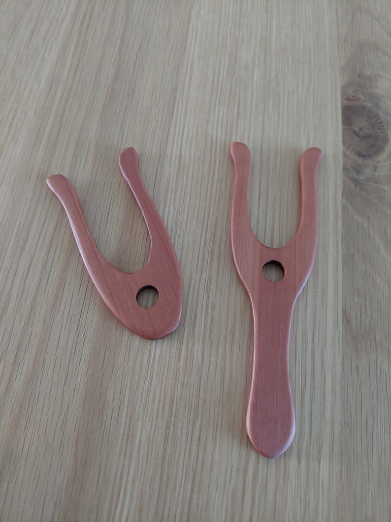 May include: Two wooden weaving tools in a reddish-brown hue, each with a distinctive U-shaped top and a central hole. One tool is smaller and more compact, while the other is longer with a handle. The tools are displayed on a light-colored wooden surface.