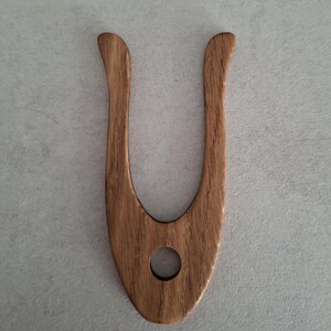 May include: A wooden hair fork with a U-shaped design. The fork is made of light brown wood with a visible grain pattern. It has a rounded hole near the base and curved tines. The hair accessory is designed for styling and securing hair.