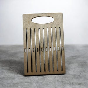 May include: A light brown wooden chopping board with a handle and vertical slots. The rectangular board has a handle at the top and a series of evenly spaced, parallel slots running down its face. The wood grain is visible.
