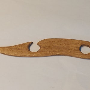 May include: A wooden knitting needle tool with a unique curved shape and a hole in the centre. The tool is made of light brown wood with a natural grain pattern.