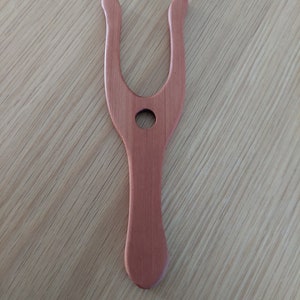 May include: A wooden weaving tool, in a light reddish-brown color, is displayed against a light wood grain surface. The tool has a Y-shaped top with rounded ends and a handle. A circular hole is in the center of the tool.