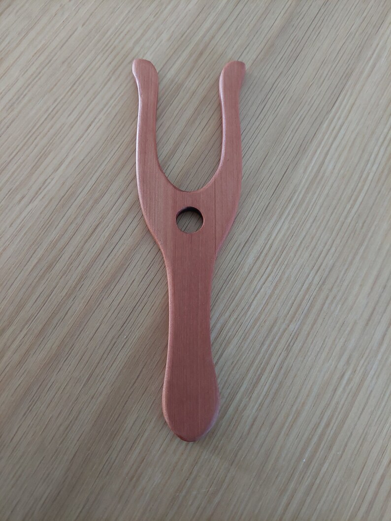 May include: A wooden weaving tool, in a light reddish-brown color, is displayed against a light wood grain surface. The tool has a Y-shaped top with rounded ends and a handle. A circular hole is in the center of the tool.
