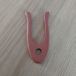 May include: A reddish-brown wooden tool with a U-shape and a circular hole in the center. The tool has a smooth finish and is likely used for crafting or woodworking. The background is a light-colored wooden surface.
