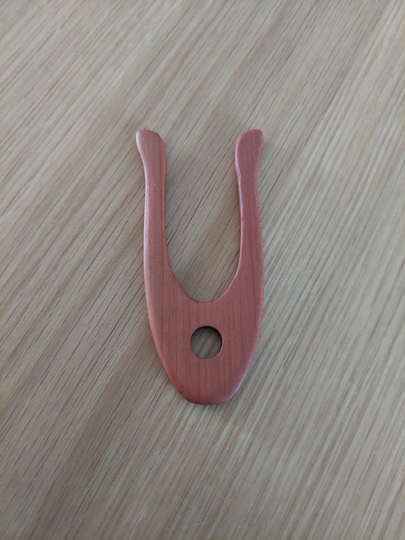 May include: A reddish-brown wooden tool with a U-shape and a circular hole in the center. The tool has a smooth finish and is likely used for crafting or woodworking. The background is a light-colored wooden surface.