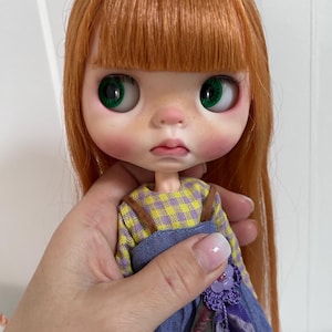 Könnte beinhalten: Eine Puppe mit langem, glattem, ingwerfarbenem Haar und Pony, großen, leuchtend grünen Augen, einer kleinen Nase und rosigen Wangen. Die Puppe trägt ein langärmeliges Hemd mit gelb-violettem Karomuster unter einem blauen Jeans-Latzkleid.