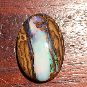 Könnte beinhalten: Ein brauner und weißer Opal-Cabochon mit einem blauen und grünen Schimmer. Der Opal ist in eine braune Matrix gefasst.