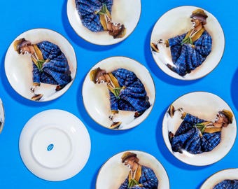 May include: Set of nine white ceramic plates with a gold rim. Each plate features a colorful illustration of a clown in a blue and white plaid suit spinning a top.