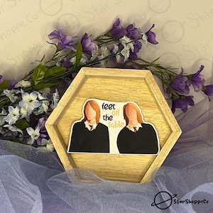 May include: A hexagonal wooden tray with a sticker of two people with brown hair wearing black robes and the text "feet off the table".