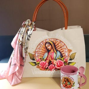 Puede incluir: Una bolsa de lona blanca con asas de cuero marr&oacute;n. La bolsa presenta una imagen colorida de la Virgen de Guadalupe con el texto "Gracias por tus bendiciones Virgen de Guadalupe". Una taza de cer&aacute;mica rosa con una cuchara y una imagen similar de la Virgen de Guadalupe est&aacute; en primer plano.