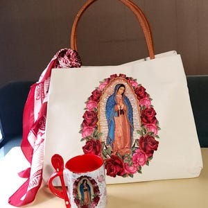 Puede incluir: Una bolsa de lona blanca con asa de cuero marr&oacute;n y una imagen colorida de la Virgen Mar&iacute;a rodeada de rosas. Una taza roja con una imagen similar est&aacute; al lado de la bolsa.