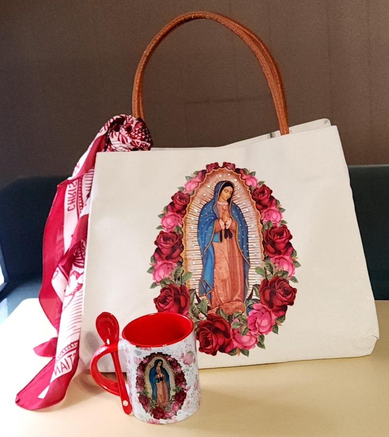 Puede incluir: Una bolsa de lona blanca con asa de cuero marr&oacute;n y una imagen colorida de la Virgen Mar&iacute;a rodeada de rosas. Una taza roja con una imagen similar est&aacute; al lado de la bolsa.