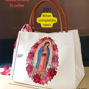 Puede incluir: Una bolsa de lona blanca con asa de cuero marr&oacute;n. La bolsa presenta una imagen colorida de la Virgen Mar&iacute;a rodeada de rosas rojas y rosas. La bolsa mide 10 pulgadas de alto, 14,25 pulgadas de ancho y 7,25 pulgadas de profundidad. Cuando est&aacute; completamente abierta, la bolsa mide 20 pulgadas de ancho.