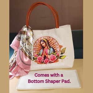 Puede incluir: Una bolsa de lona blanca con asa de cuero marr&oacute;n y una imagen colorida de la Virgen de Guadalupe. La bolsa tiene el texto "MAS POR TUS BENDICIONES VIRGEN DE GUADALUPE" impreso en ella. La bolsa viene con una almohadilla de forma de fondo de lona blanca.