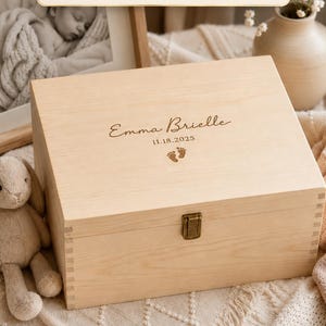 May include: A rectangular light-colored wooden keepsake box with a metal latch. The lid is engraved with the name Emma Brielle, the date 11.18.2025, and a small footprint icon. Text on the image reads 14 Designs - Weddings, Baby, Pets, Memorial, Baptism, & More, Solid Wood, and US-Crafted.
