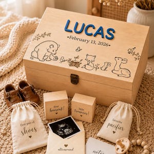 May include: A wooden keepsake box with the name LUCAS and February 13, 2026 in blue and black text, featuring woodland animal engravings. Includes two small wooden blocks, two drawstring bags, an ultrasound envelope, and baby shoes.
