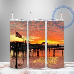 Puede incluir: Tres vasos de acero inoxidable con una escena de puesta de sol sobre el agua. El vaso de la derecha tiene un contorno dorado del estado de Carolina del Norte con la palabra "Bienvenido" en su interior.