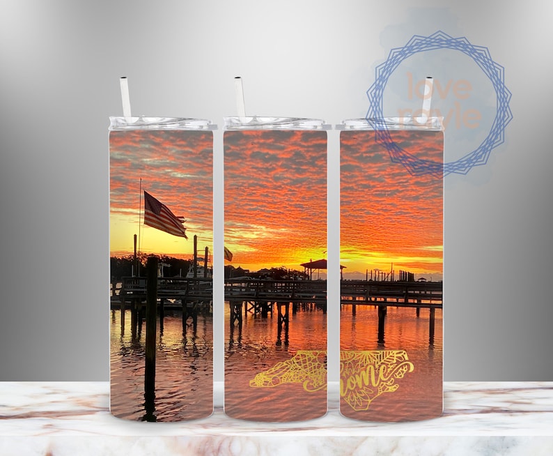 Puede incluir: Tres vasos de acero inoxidable con una escena de puesta de sol sobre el agua. El vaso de la derecha tiene un contorno dorado del estado de Carolina del Norte con la palabra "Bienvenido" en su interior.