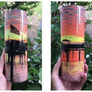 Puede incluir: Un vaso de acero inoxidable con una escena de puesta de sol y un muelle con una silueta de un edificio y una bandera. La parte inferior del vaso tiene la palabra "home" en una escritura amarilla.