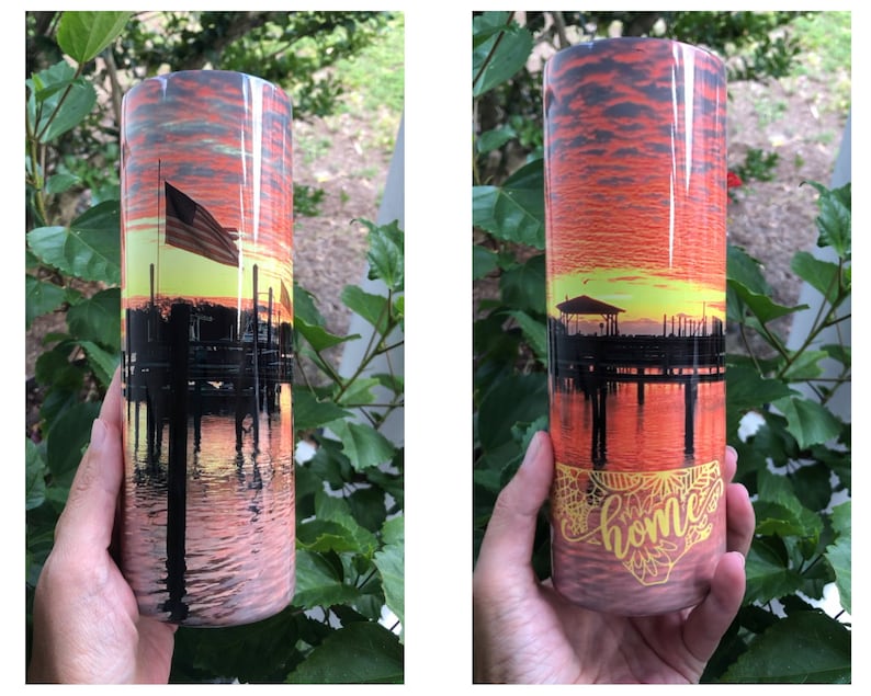 Puede incluir: Un vaso de acero inoxidable con una escena de puesta de sol y un muelle con una silueta de un edificio y una bandera. La parte inferior del vaso tiene la palabra "home" en una escritura amarilla.