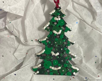 May include: A green Christmas tree ornament with white snow and glitter accents. The ornament is hanging from a red ribbon.