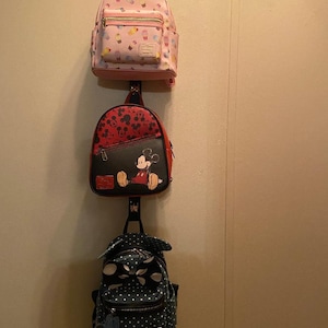 May include: Three mini backpacks hanging on a hook. The top backpack is pink with a cupcake pattern. The middle backpack is red with a Mickey Mouse pattern. The bottom backpack is blue with white polka dots and a Mickey Mouse charm.