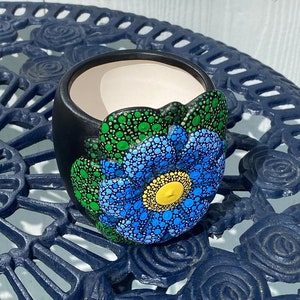 May include: A black ceramic planter with a hand-painted blue, green, and yellow flower design. The planter is decorated with a dot painting technique.