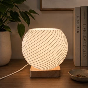 May include: A spherical table lamp with a white, ribbed, spiraling texture that emits a soft glow. The lamp is mounted on a square, light-colored wooden base and includes a white power cord extending from the side of the base.